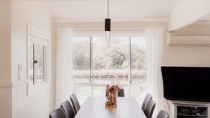 A conference room with a long wooden table, black chairs, a hanging light fixture, and a large window with a view of trees.