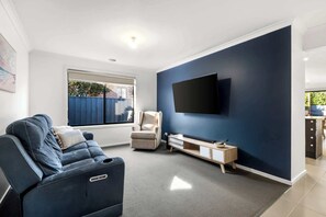 Een moderne woonkamer met een donkerblauwe accentmuur, een flatscreen televisie boven een houten dressoir, een grijze bank met een kussen, en een raam met uitzicht op een hek en groen.