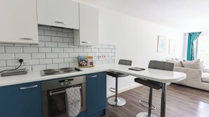 A modern kitchen with white cabinets, a built-in oven, and a white countertop. There is a dining area with a white table and two bar stools. - West Hull 2 bed apartment with Parking (Hull)