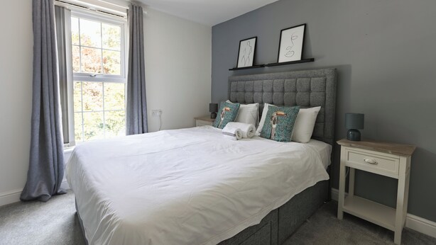 A bedroom with a large bed, grey curtains, a bedside table with a lamp, and two framed pictures on the wall. - West Hull 2 bed apartment with Parking (Hull)