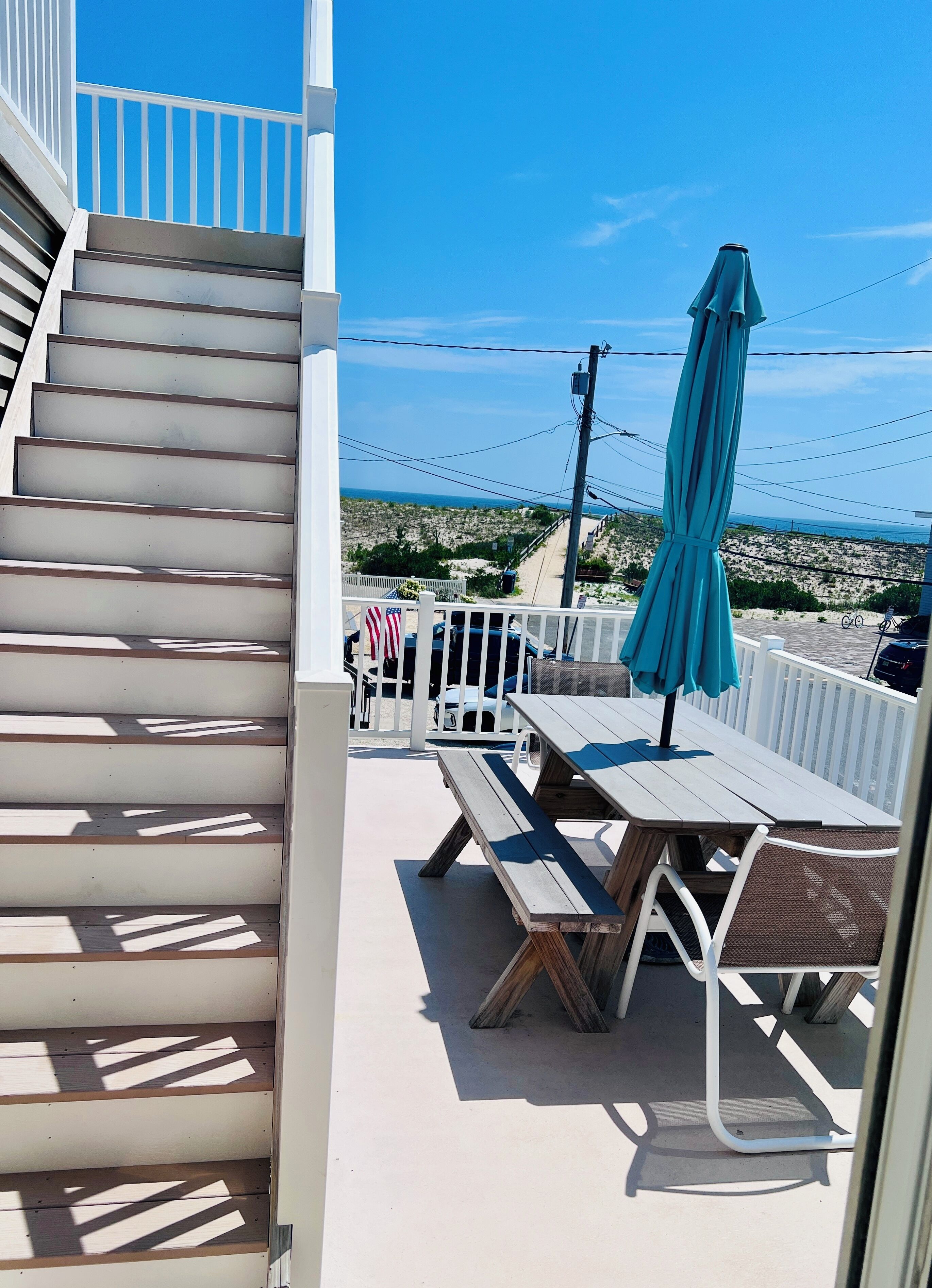 A balcony with a wooden table and chairs, a turquoise umbrella, and a view of the beach and ocean.