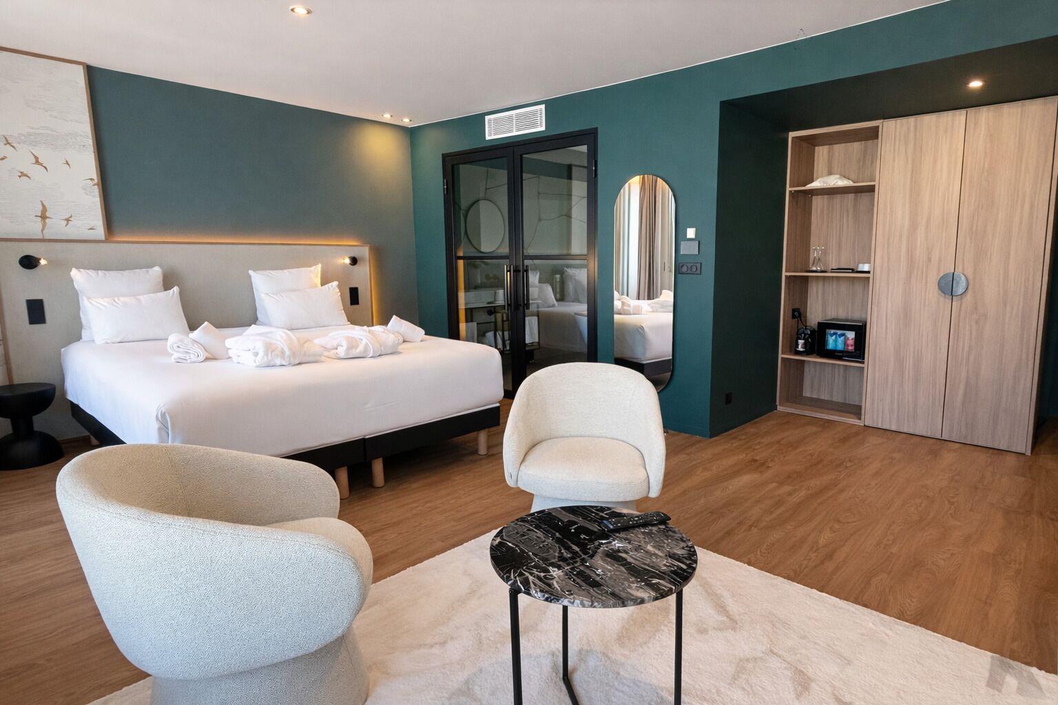 A modern hotel room with a bed, two chairs, a small table, a wardrobe, and a mirror.