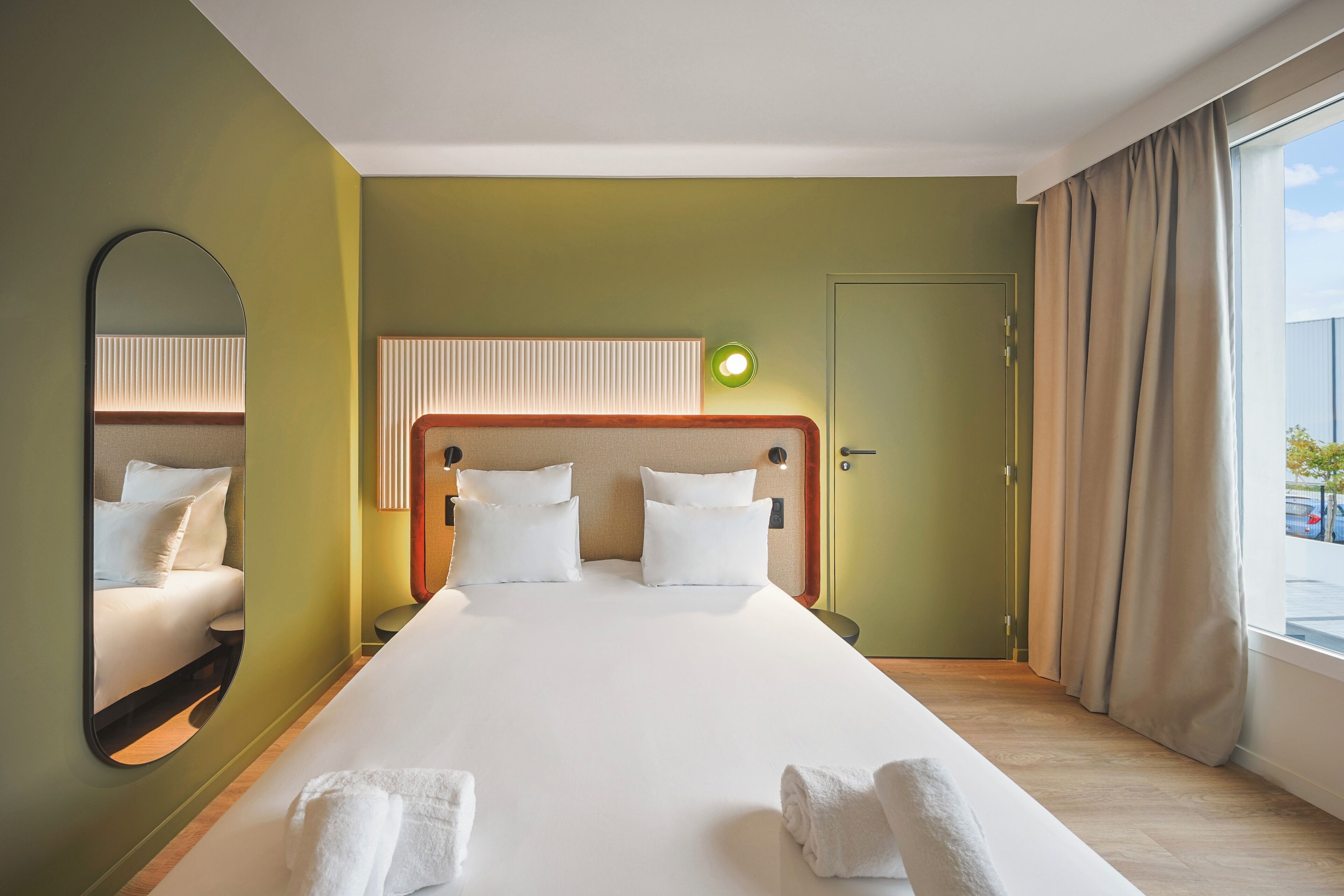 A hotel room with a large bed, a mirror, a bedside table, and a window with curtains.