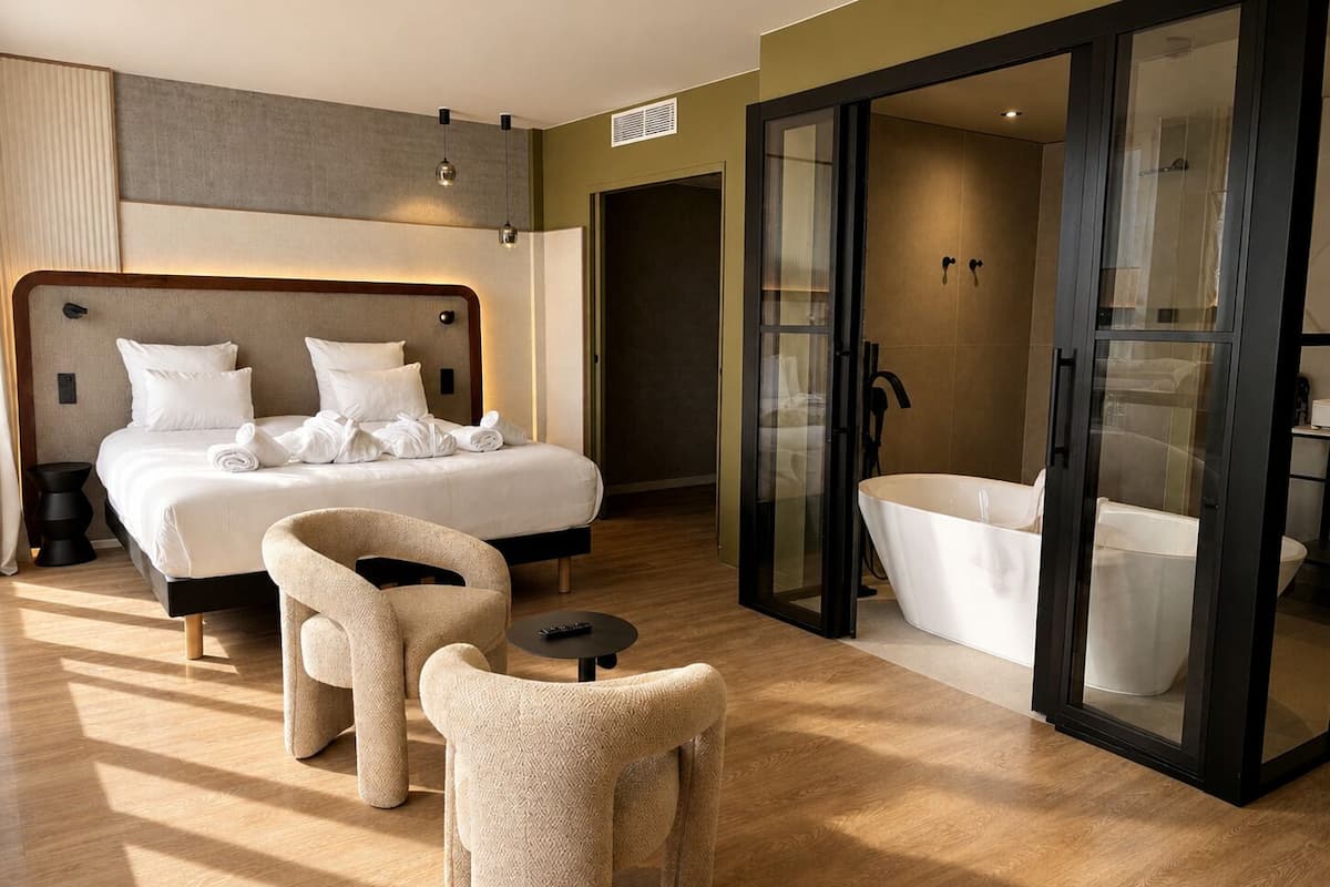 Une chambre d’hôtel moderne dotée d’un grand lit, d’une salle de bain séparée avec une douche en verre, et d’un coin salon avec deux fauteuils.