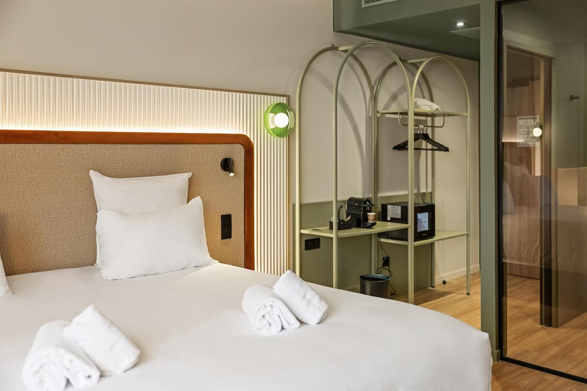 Une chambre d’hôtel avec un lit bien fait, une tête de lit dotée d’un luminaire, un petit bureau avec un ordinateur et une cabine de douche en verre.