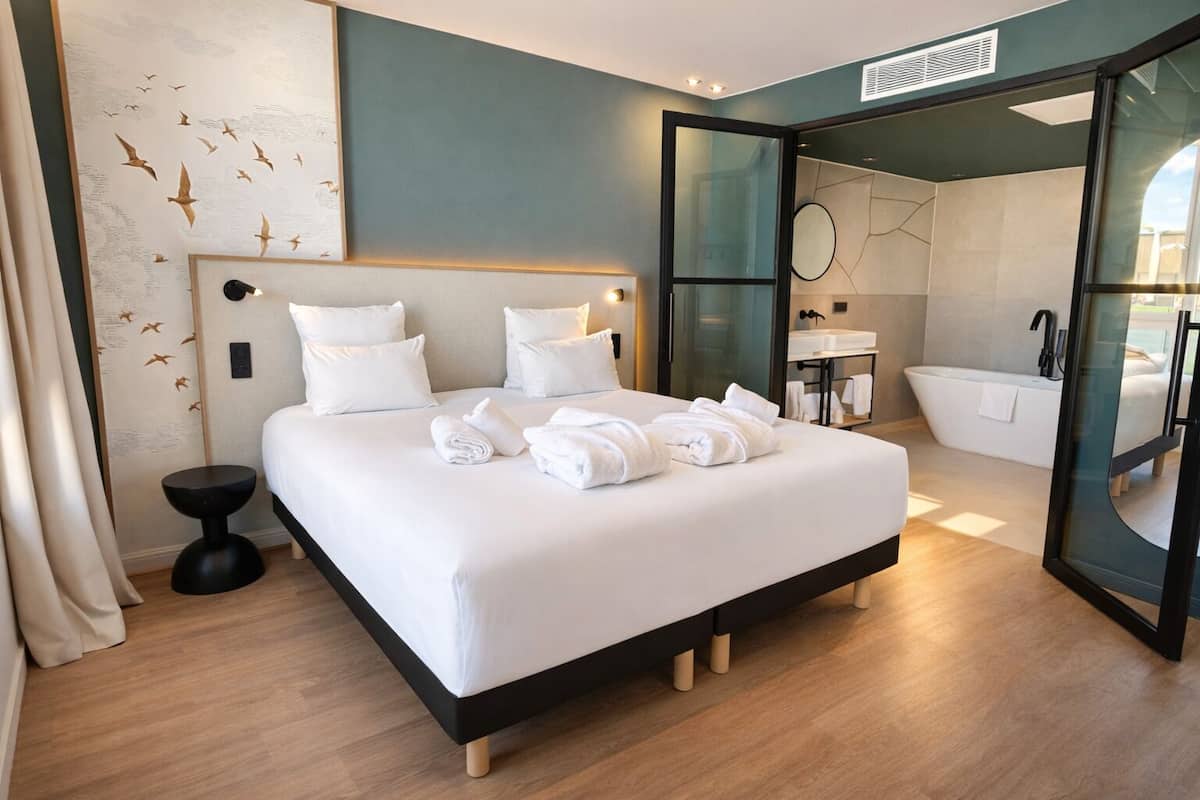 Une chambre d’hôtel moderne dotée d’un grand lit, d’une salle de bain avec baignoire et d’une porte en verre donnant accès à la salle de bain.