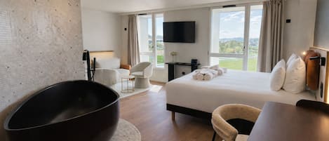 A modern hotel room with a large bathtub, a bed with neatly folded towels, a flat-screen TV, and a view of the outdoors.