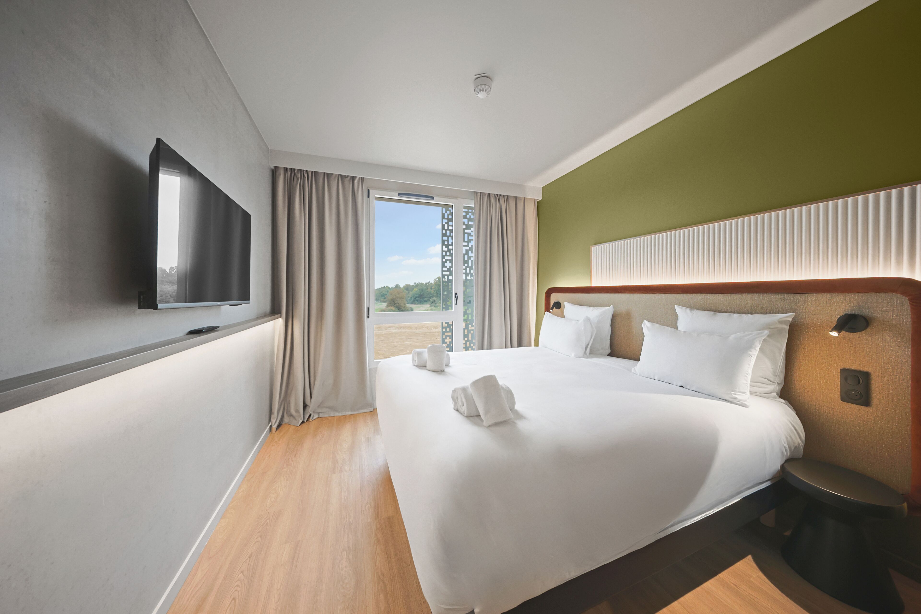 Quarto de hotel com uma cama grande, uma televisão de tela plana na parede, piso de madeira e vista através de uma porta de vidro.
