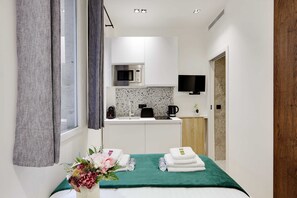 Una habitación de hotel pequeña con una cama, una manta verde, un ramo de flores y una kitchenette compacta con microondas y cafetera.