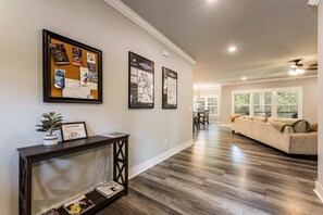 A hallway with framed informational posters, a console table with a potted plant, and a living area with a sofa and windows. - The Abode in Bella Vista .7mi to Back 40 Trailhead (Bella Vista)