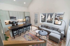 Une cuisine moderne avec un Ăźlot central, un coin repas avec des chaises, un canapĂ© gris et une grande fenĂȘtre donnant sur des maisons.