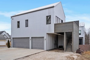 Un edificio moderno de estilo industrial con dos puertas de garaje, un acceso cubierto con escaleras y una estructura adyacente más pequeña que dice 'Entrada al Garaje'.