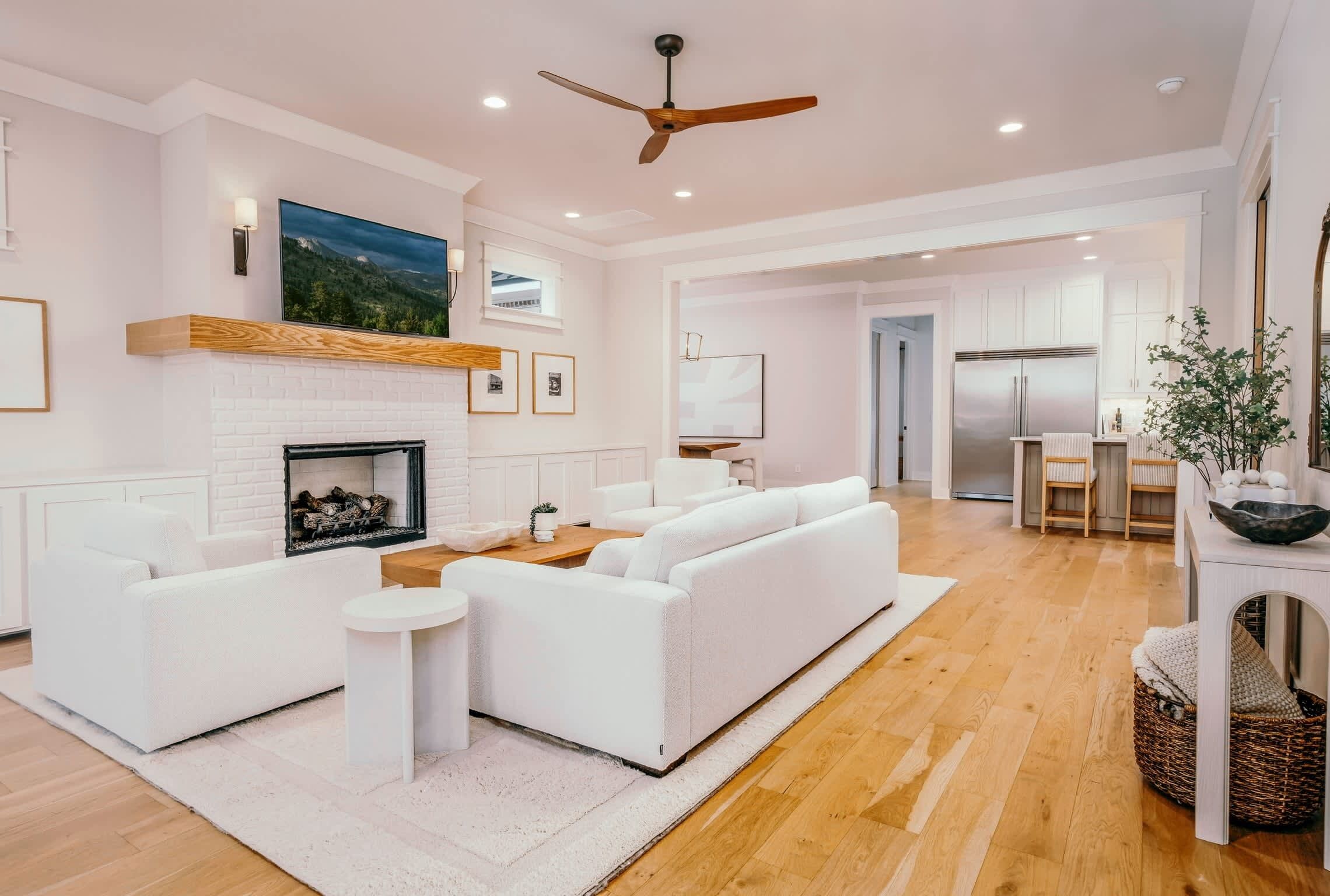 A spacious living room with a white fireplace, a ceiling fan, and a kitchen area in the background.
