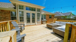 Une terrasse en bois avec des meubles d’extérieur, un patio couvert et une maison en briques dotée de grandes fenêtres.