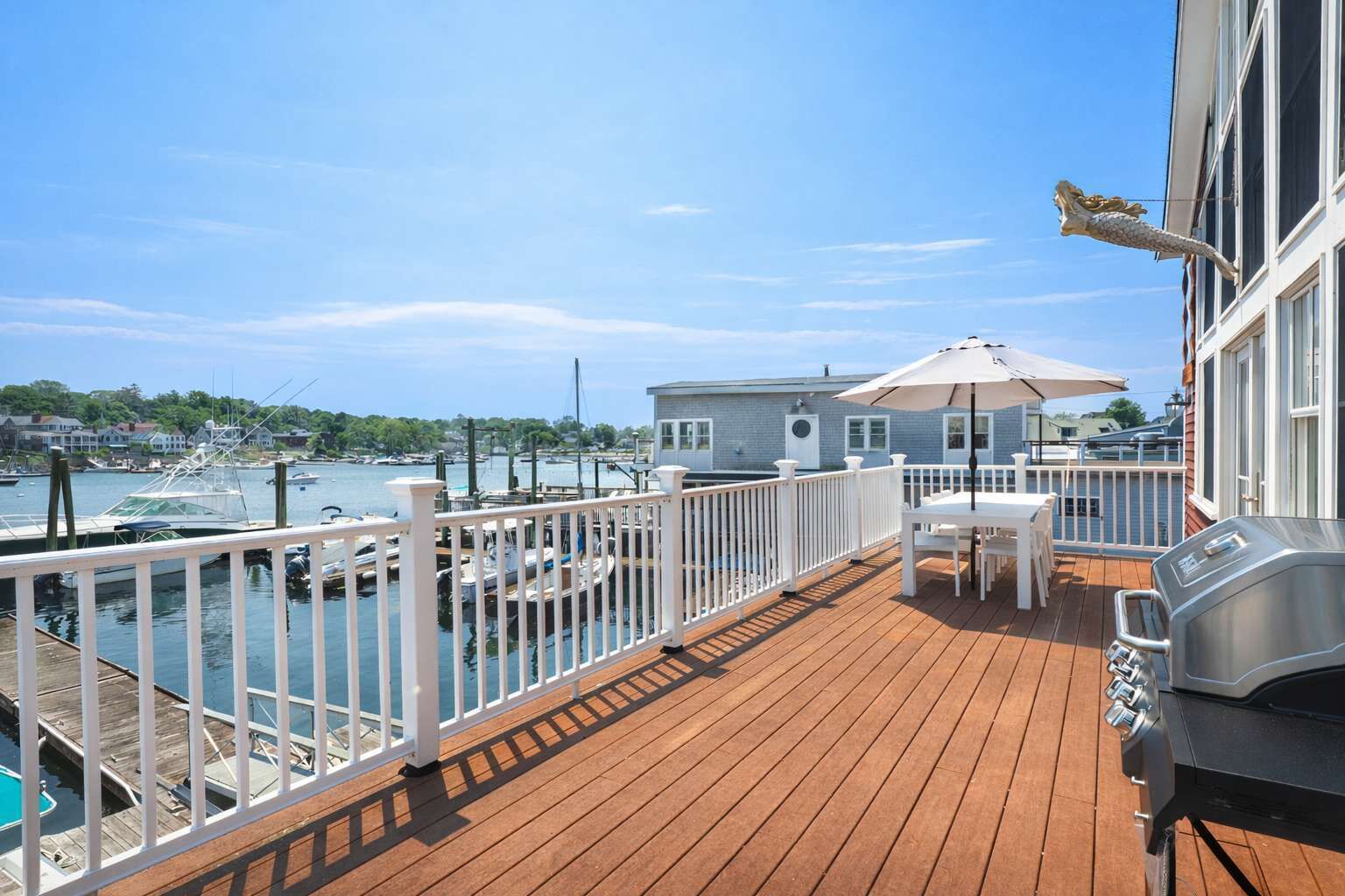 Een houten terras met een barbecue, een tafel met parasol en uitzicht op een jachthaven met boten.