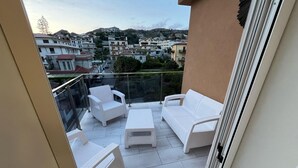 A balcony with white wicker furniture, including two chairs and a sofa, overlooking a residential area with houses and hills. - Rupella  (Roccella Jonica)