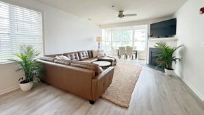 A modern living room with a brown leather sofa, a coffee table, and a fireplace. There is a dining area with a table and chairs visible through large windows. - Condo with Community Pool - 17432 Slipper Shell Way Unit 1 (Lewes)
