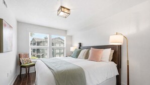 Un dormitorio con una cama grande, una mesita de noche, una lámpara de pie, un sillón con cojín y una ventana con persianas.