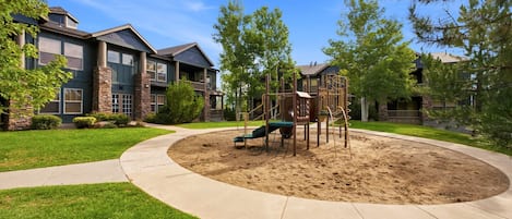 Un parque infantil con tobogán y estructuras para escalar, rodeado de una zona ajardinada con un sendero curvo.