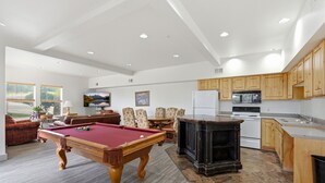 Amplia sala de estar con mesa de billar, asientos cómodos, cocina con gabinetes de madera y un ventanal con vistas al exterior.