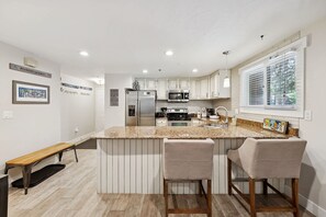 A modern kitchen with white cabinetry, a granite countertop, a stainless steel refrigerator, and a microwave. There are two bar stools and a bench.