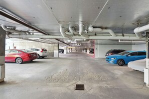 Un garaje para estacionamiento con varios coches aparcados, suelo de hormigón y tuberías visibles en el techo.