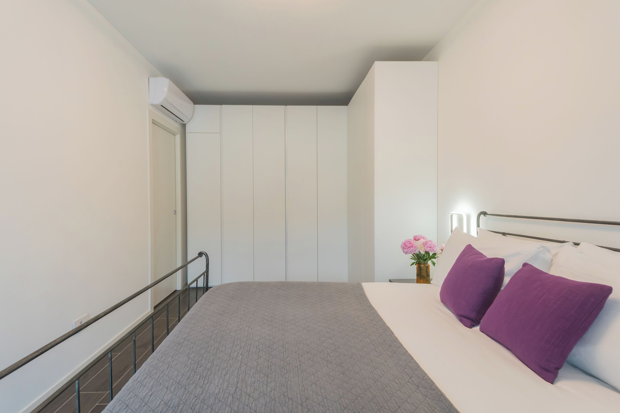 A bedroom with a bed, a nightstand, a vase with flowers, and a wall-mounted light.