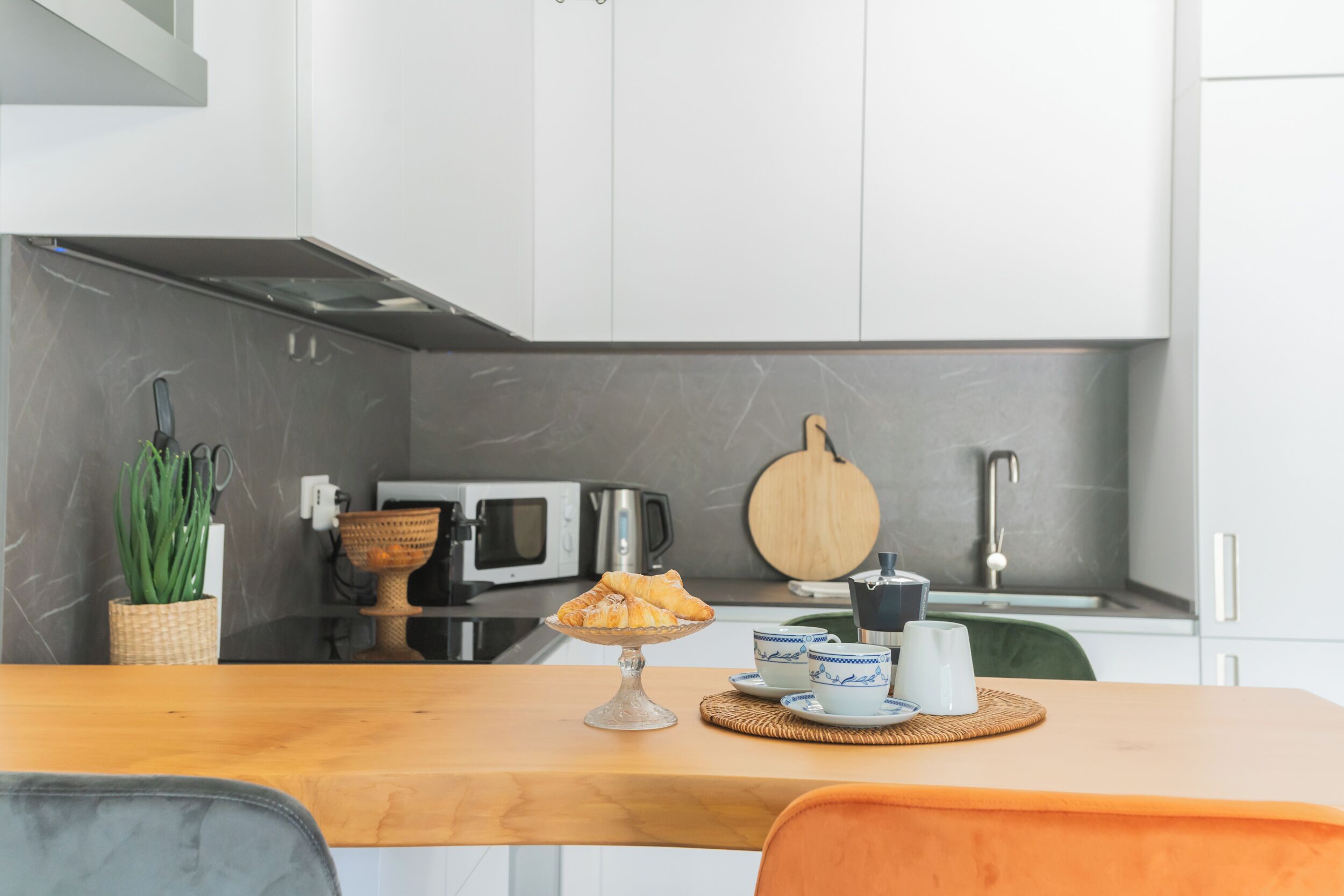 A modern kitchen with white cabinets, a wooden countertop, a microwave, a coffee maker, and a plate of croissants.