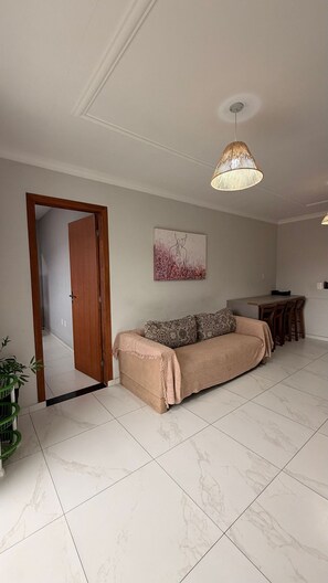 A spacious living area with a sofa, a dining table with chairs, a hanging light fixture, and a door leading to another room. - Residencial Águia - Piske Imóveis (Florianopolis)