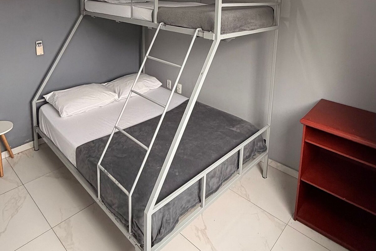 Un lit superposé avec une échelle, une petite table et une étagère rouge dans une pièce aux murs gris et au plafond blanc.