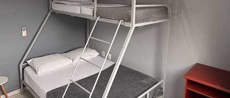Una cama litera con escalera, una mesita pequeña y un estante rojo en una habitación con paredes grises y techo blanco.