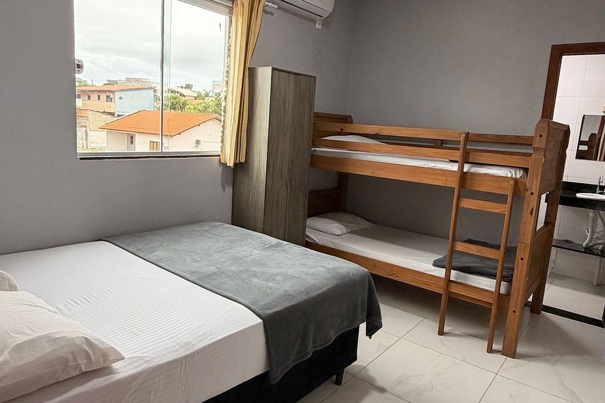 A bedroom with a bunk bed, a desk, a chair, a window with curtains, an air conditioning unit, and a view of buildings outside.