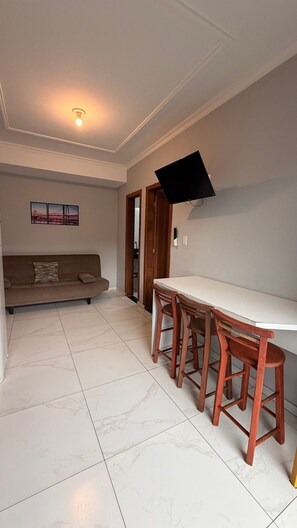 Ein Wohnzimmer mit einer Couch, einem Esstisch mit Stühlen, einem an der Wand befestigten Flachbildfernseher und einer Deckenleuchte.