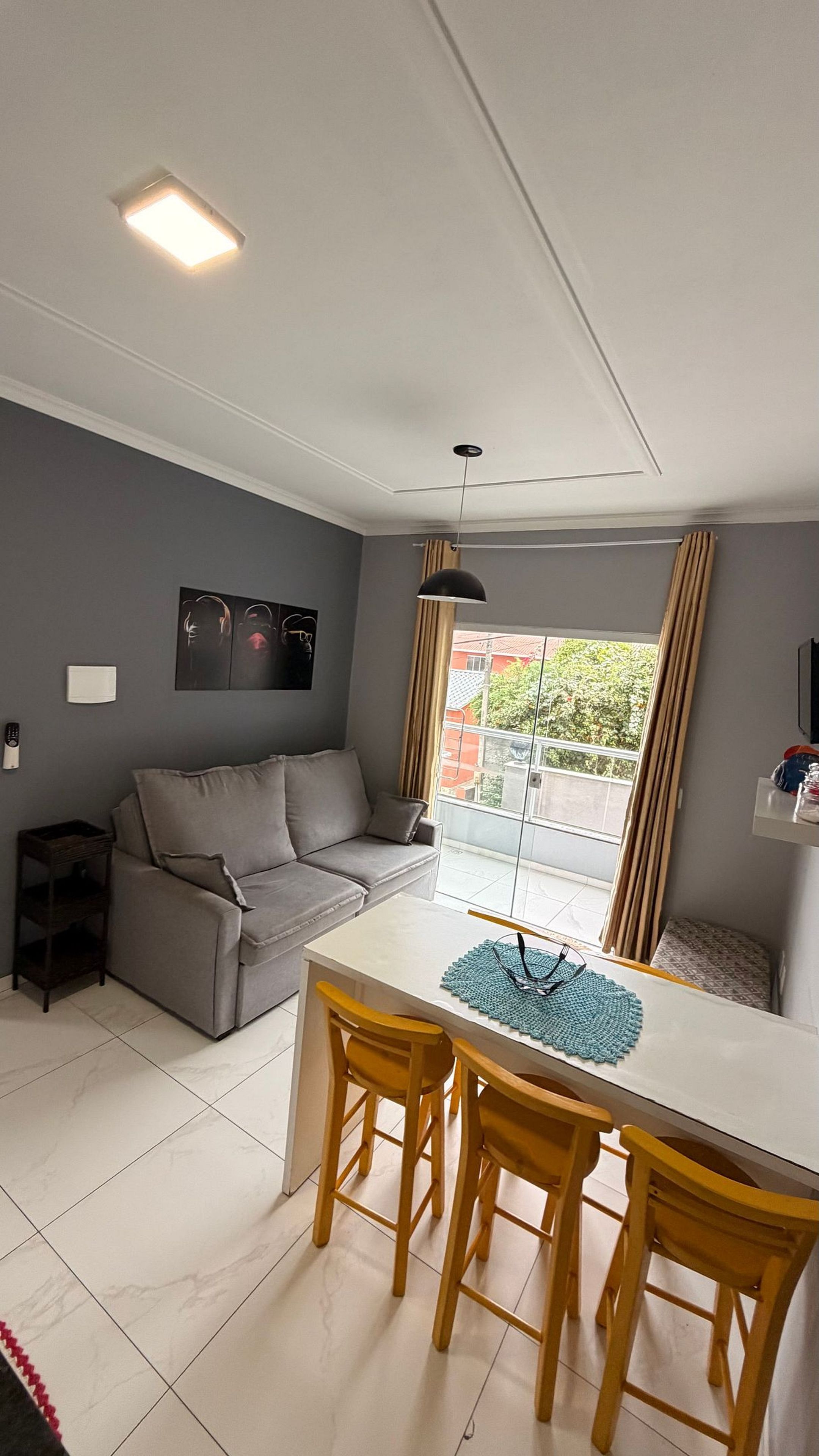Uma sala de estar moderna com um sofá cinzento, uma mesa de jantar branca com cadeiras amarelas, uma luminária no teto e vista para uma varanda.