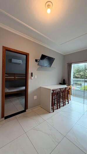 A room with a bunk bed, a small kitchenette, a flat-screen TV, a dining area with chairs, and a large window. - Residencial Águia - Piske Imóveis (Florianopolis)