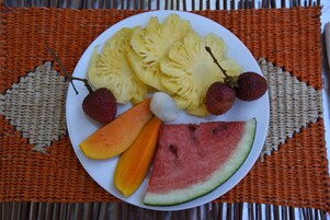Un plato de fruta con piña, mango, papaya, sandía y lichi sobre una base tejida.