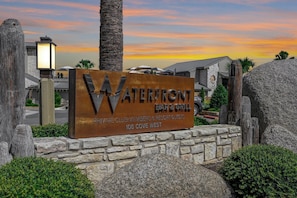 A sign for Waterfront Bar & Grill with a stone wall, palm trees, and a sunset in the background.