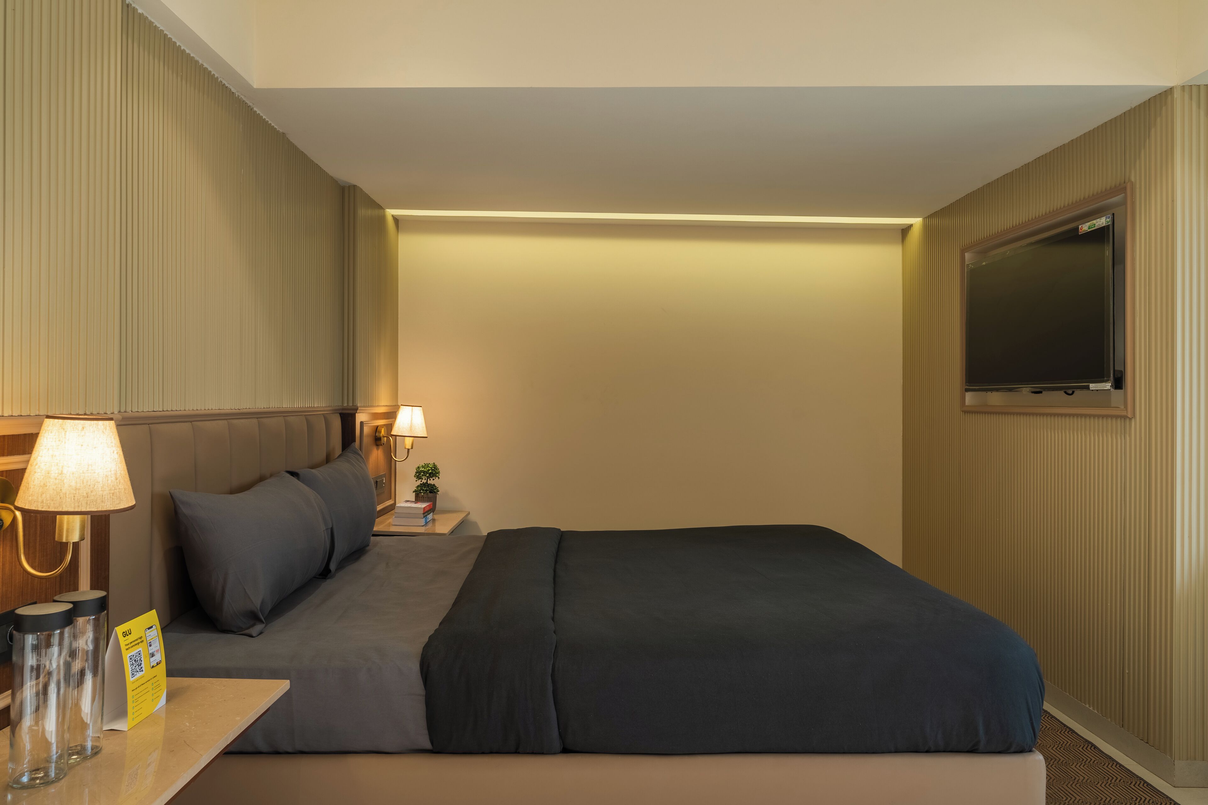 A hotel room with a bed, bedside lamp, a small table with a lamp, a TV, and a sign on the table.
