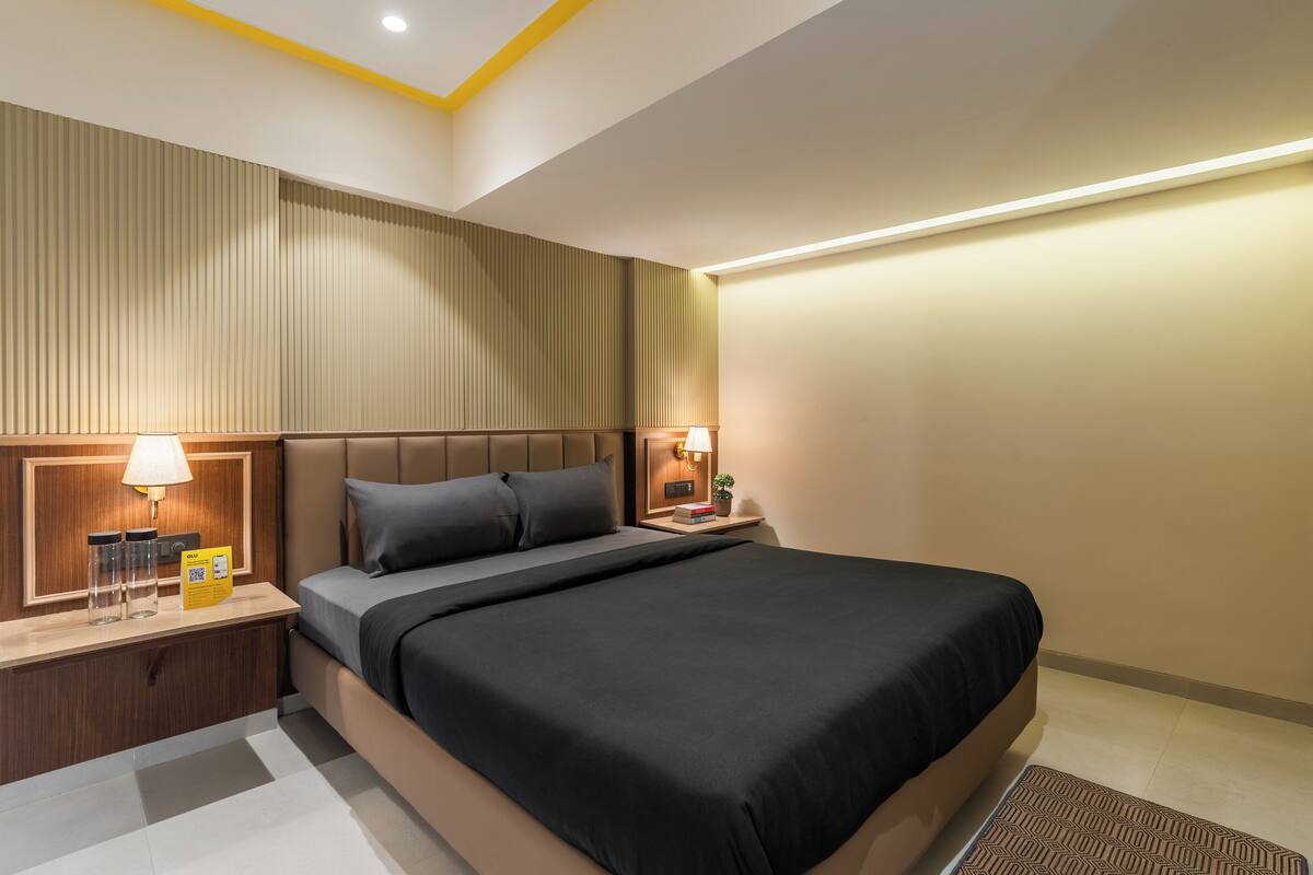 A modern hotel room with a large bed, a nightstand with a lamp, a small table with a plant, and a wall-mounted panel.
