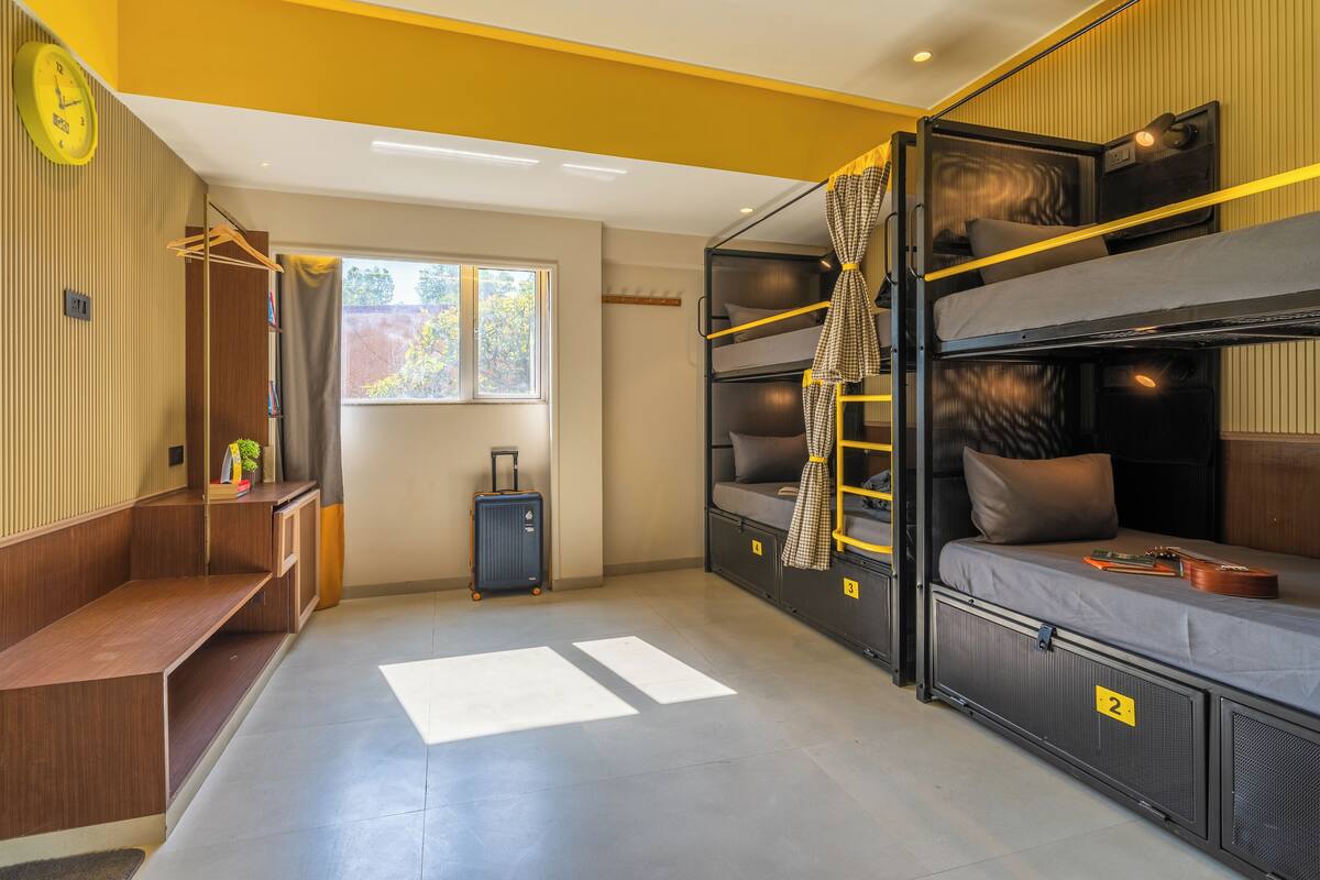 A compact room with bunk beds, a small desk, a suitcase, and a window with a view of trees.