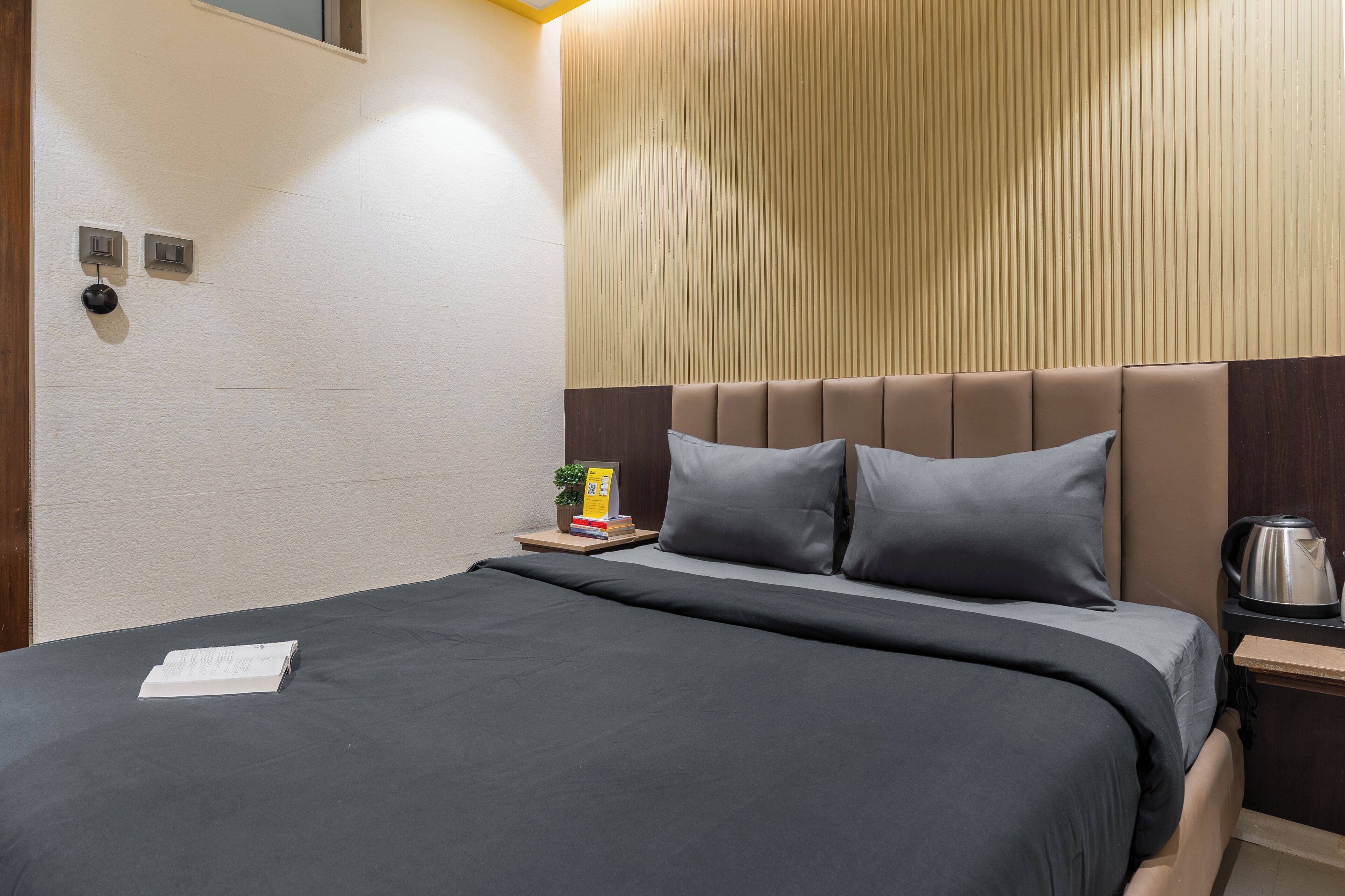 A neatly made bed with a dark bedspread, a wooden headboard, and a nightstand with a kettle and books.