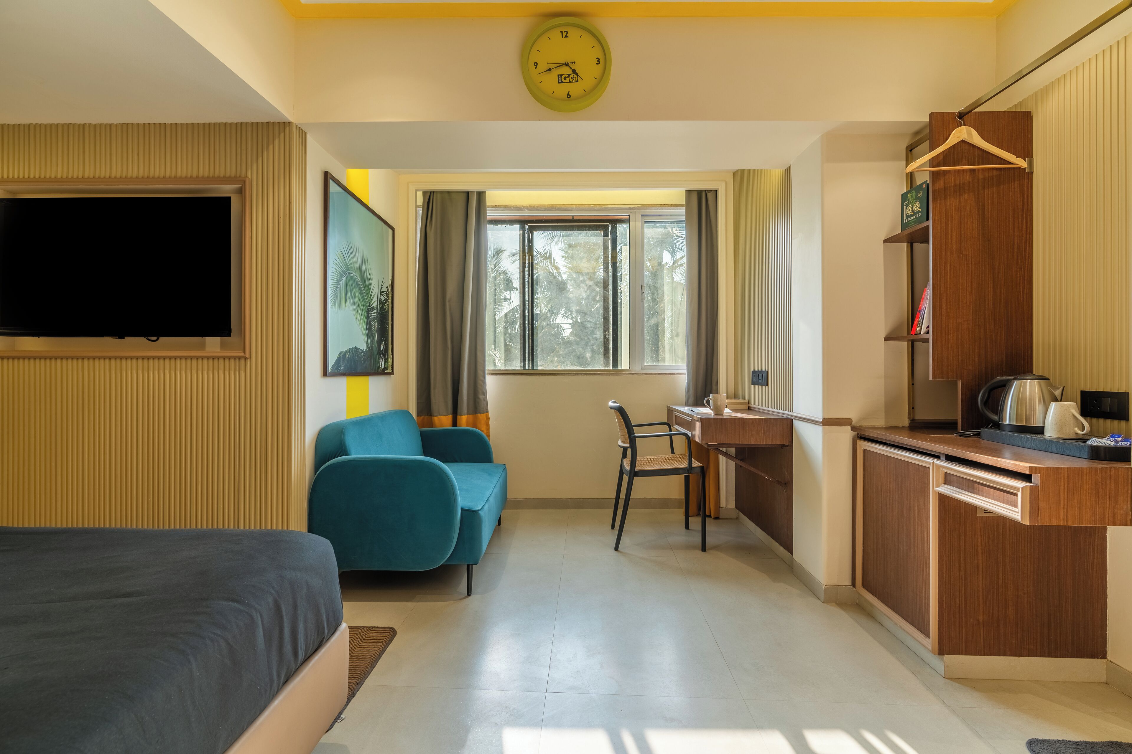 Una camera d'hotel con un letto, una scrivania, una sedia, una televisione, una finestra con tende e una sveglia appesa al muro.
