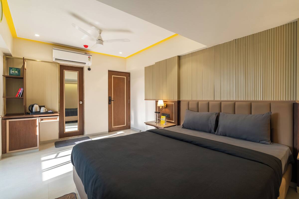 A modern hotel room with a large bed, a desk with a chair, a wardrobe, and a bedside table with a lamp.