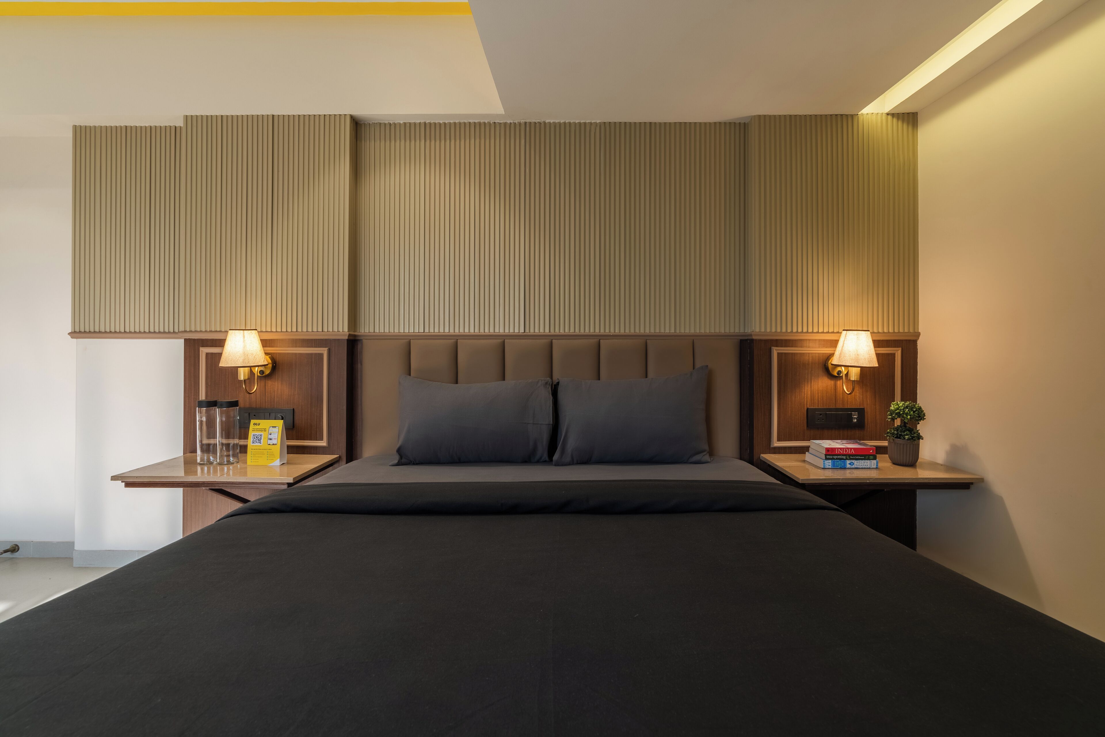 A modern hotel room with a large bed, two bedside tables with lamps, a small plant, and a nightstand with a book and a glass.
