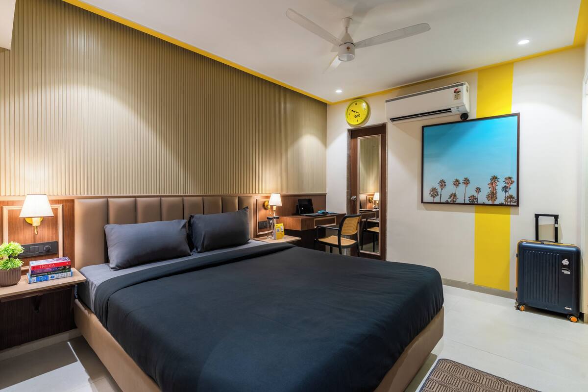 A modern hotel room with a large bed, bedside table with a lamp, a desk with a chair, a ceiling fan, an air conditioning unit, and a painting on the wall.