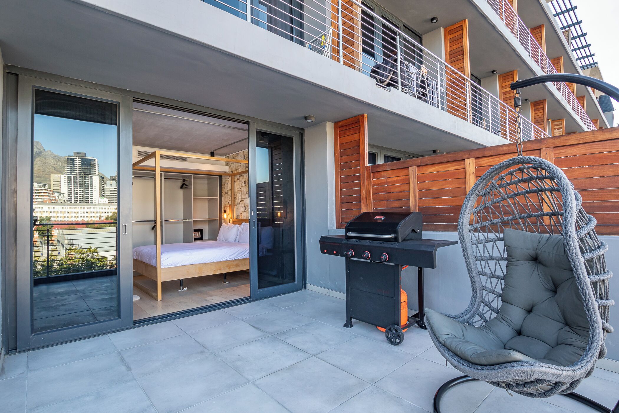 Ein moderner Balkon mit Grill, Hängesessel und einem Schlafzimmer mit Bett und Kleiderschrank.