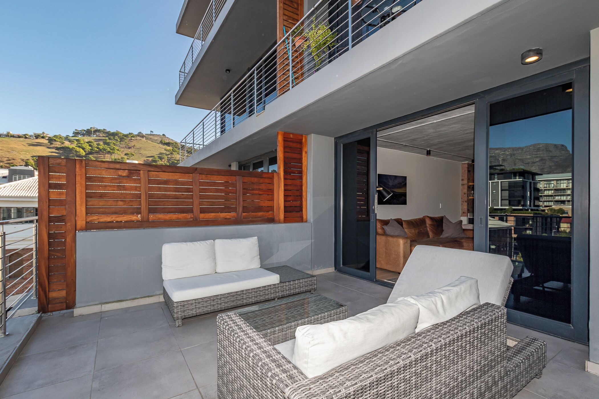 A modern outdoor living area with wicker furniture and a wooden fence, overlooking a hilly landscape.