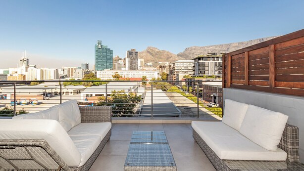 Una terrazza sul tetto con un divano bianco e un tavolo in vetro, con vista su un paesaggio urbano e montagne sullo sfondo.