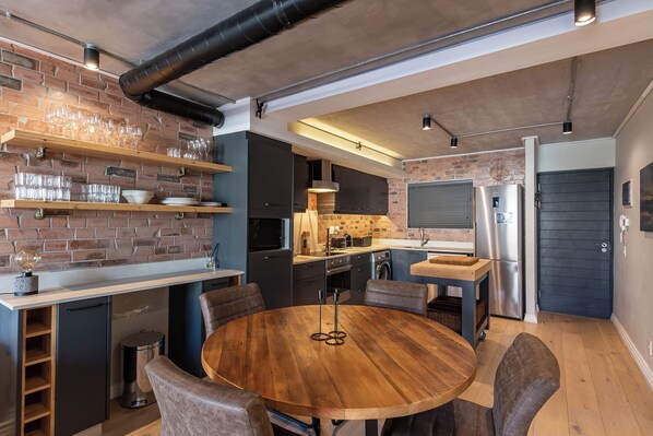 A modern kitchen with brick walls, stainless steel appliances, and a round wooden dining table. - The Docklands Two Bed Superior Apartment (Cape Town)