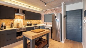 Una cucina moderna con mobili scuri, una parete decorata con mattoni, un tavolo di legno ed elettrodomestici in acciaio inossidabile.
